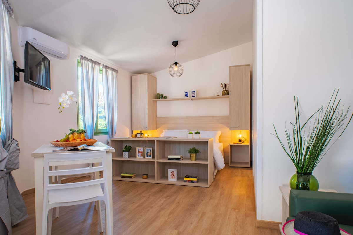 A modern, well-lit room with a wooden floor, a white desk with a basket of fruit, a green chair, and a bed with a headboard.