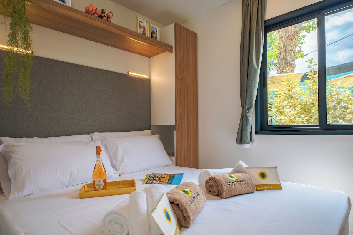 A neatly made bed with white linens, a bottle of wine, a glass, and towels with a logo.