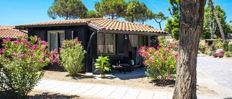 Una pequeña casa de una sola planta con tejado de tejas, un patio con mobiliario exterior y rodeada de plantas y árboles.