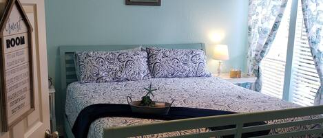 A bedroom with a bed, a ceiling fan, a framed sign, and a window with patterned curtains.