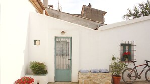 Un bâtiment aux murs blancs, avec une porte verte, un pot de fleurs, un vélo et une fenêtre ornée d’une grille métallique.