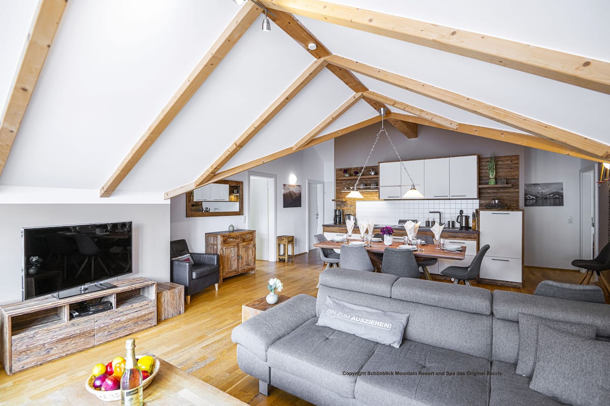 A spacious loft with a high ceiling, wooden beams, a grey sofa, a dining area, and a kitchen.
