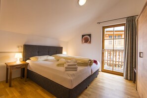 Una cama bien tendida con almohadas y una manta, una mesita de noche con una lámpara, y una ventana con vistas a edificios.