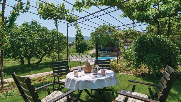 A covered outdoor seating area with a table and chairs, surrounded by greenery and a pool in the background. - Sunflower House With a Pool and Large Garden (Omis)