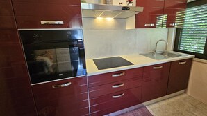 A modern kitchen with red cabinetry, a built-in oven, a stainless steel range hood, and a window with blinds. - Sunflower House With a Pool and Large Garden (Omis)