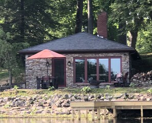 A small stone cabin with a red roof, large windows, and a wooden deck. - Hidden Cove (Findley Lake)