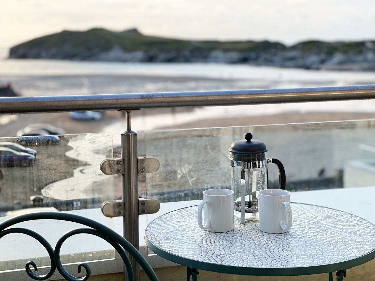 A balcony with a table set for two, a French press, and a view of the beach and sea.