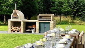 An outdoor dining area with a stone barbecue and a long table set for a meal. - Muir Estate (Stirling)
