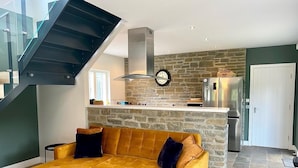 A modern living room with a mustard-colored sofa, a kitchen with a stone wall, and a staircase. - Muir Estate (Stirling)
