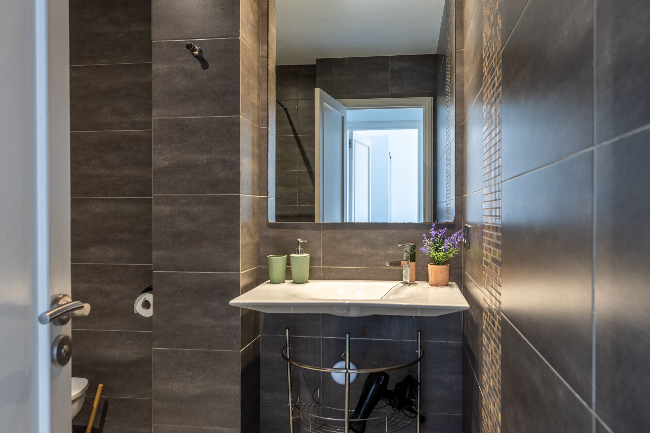 A modern bathroom with a large mirror, dark tiles, a white sink, and a shower area.