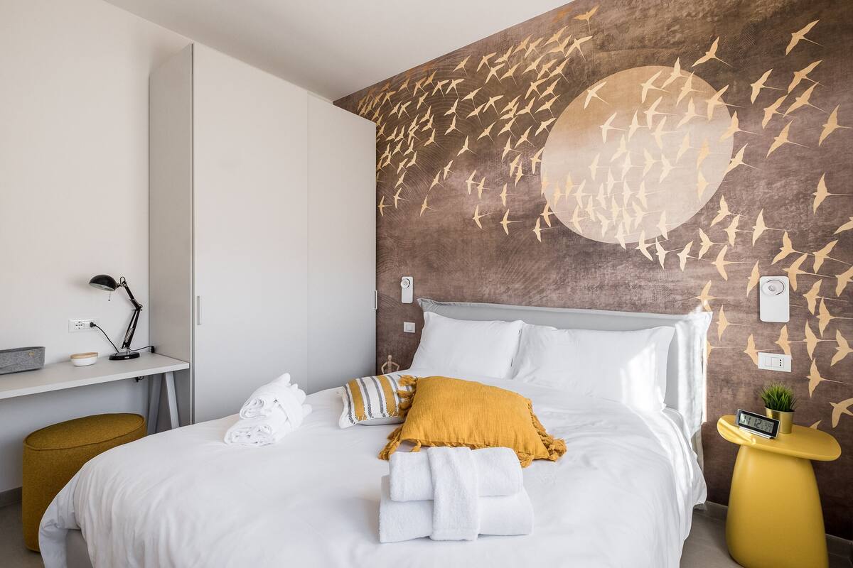 A neatly made bed with white and mustard pillows, a bedside table with a clock, and a wall with a bird pattern.
