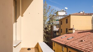 Un balcone con un piccolo tavolo di legno e una ciotola di metallo, con vista su una strada con edifici dalle tegole di terracotta e un albero.