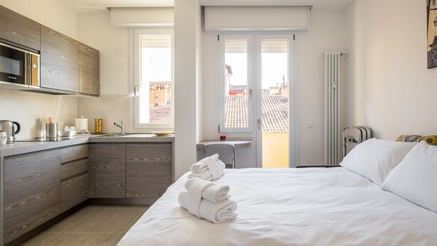 Camera d'hotel moderna con angolo cottura, un letto e asciugamani ripiegati con cura, e vista sulla città attraverso ampie finestre.