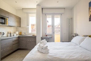 A modern hotel room with a kitchen area, a bed with neatly folded towels, and a view of the city through large windows. - Residenza del Reno by Wonderful Italy (Bologna)