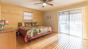 A bedroom with a wooden floor, a bed with a patterned bedspread, a ceiling fan, a window with blinds, and a view of trees through a sliding glass door. - Cabin Sleeps 12, Hot Tub, Movie Room, Game Room (Sevierville)