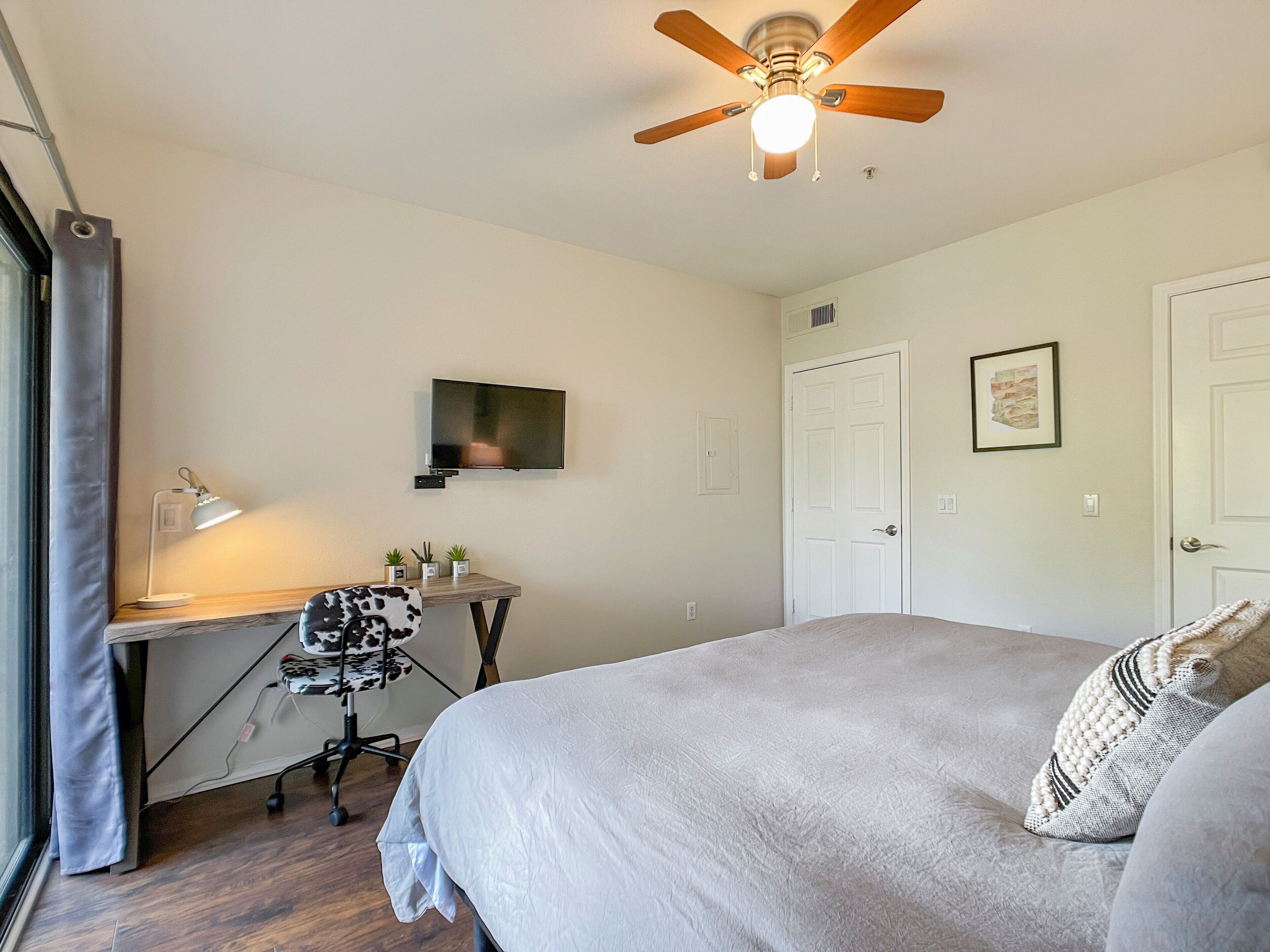 A bedroom with a bed, a desk with a computer, a chair, a ceiling fan, and a TV on the wall.