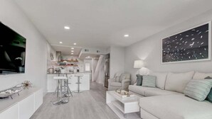 A modern living room with a sofa, a coffee table, and a TV. There is a kitchen area with a dining table and chairs.