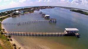 A coastal area with a long wooden pier extending into calm waters, surrounded by a sandy beach and a small structure at the end.