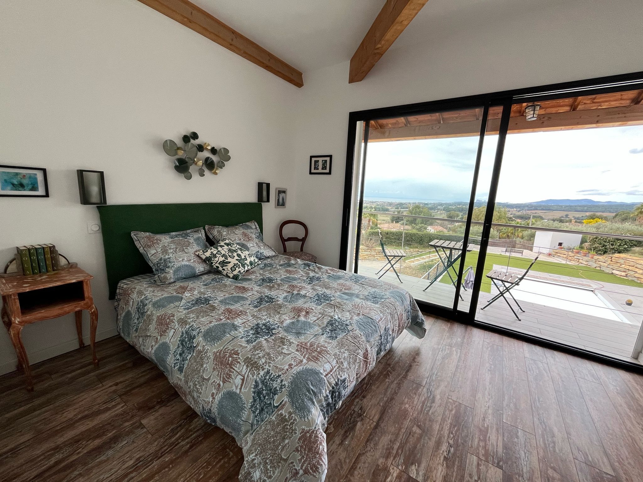 Una camera da letto con un letto grande, pavimento in legno, una testiera verde, vista sull'esterno tramite una porta scorrevole in vetro e un tavolino con libri.
