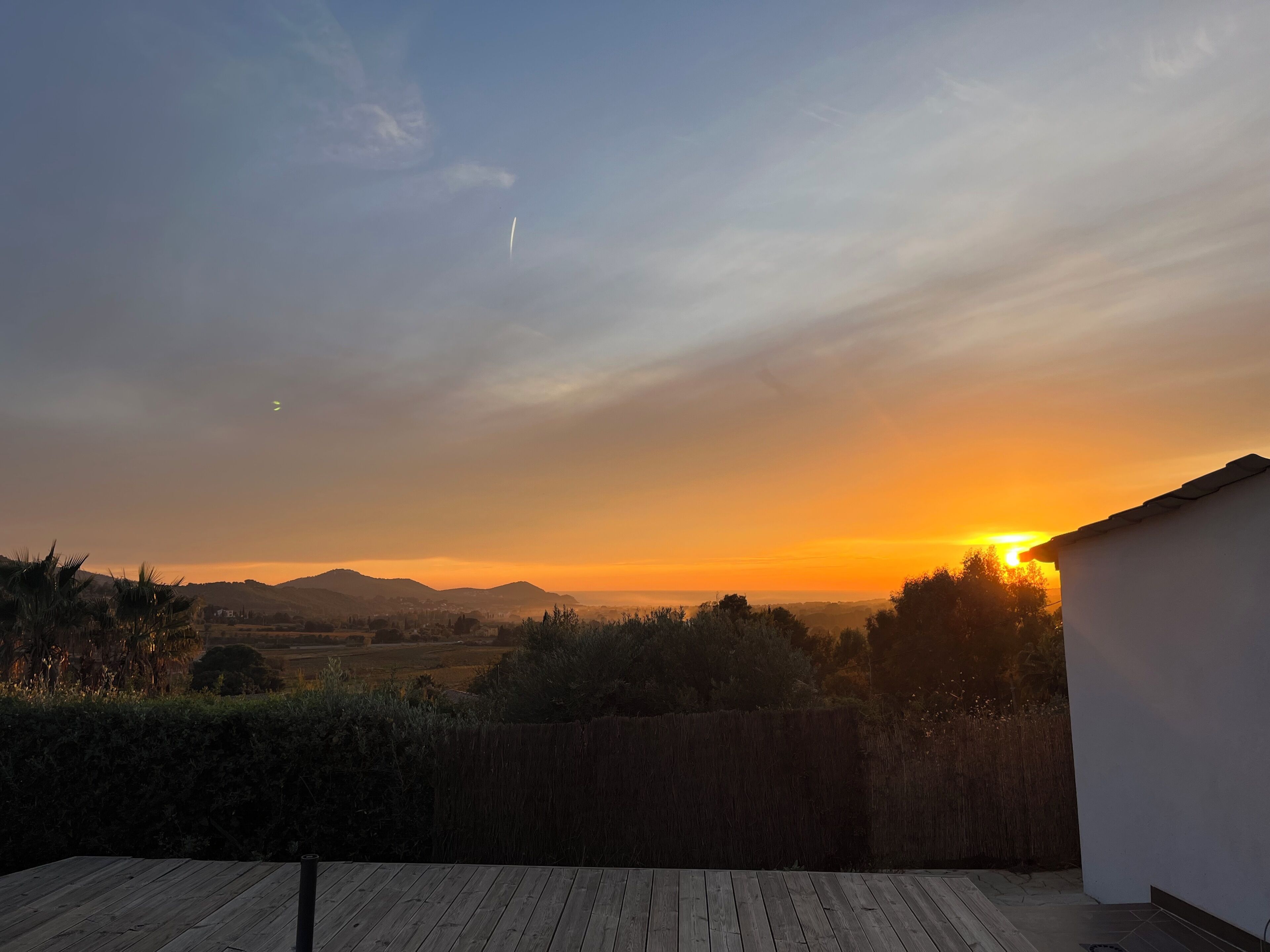 Une vue sur le coucher de soleil depuis un balcon doté d’une terrasse en bois, donnant sur un paysage rural avec des collines et un ciel dégagé.