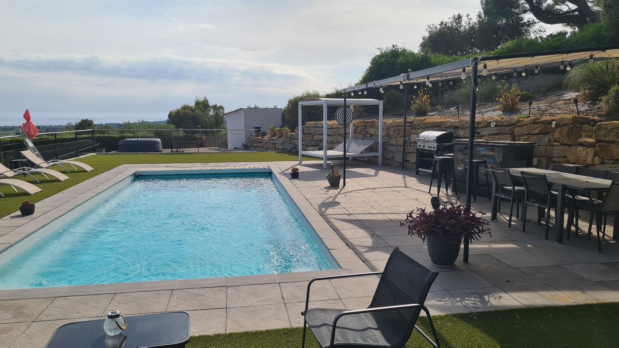 A rectangular swimming pool with a stone deck surrounding it, outdoor seating, and a barbecue area.