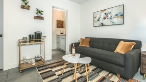 Ein modernes Wohnzimmer mit einem dunkelgrauen Sofa, einem kleinen weißen Tisch und einem gemusterten Teppich. Durch einen offenen Durchgang ist eine Küche zu sehen.
