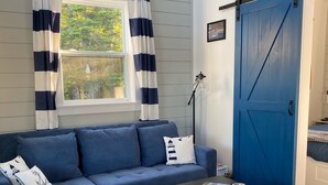 Un salon comprenant un canapé d’angle, une petite table, un fauteuil gris, une porte bleue et une fenêtre avec des rideaux.