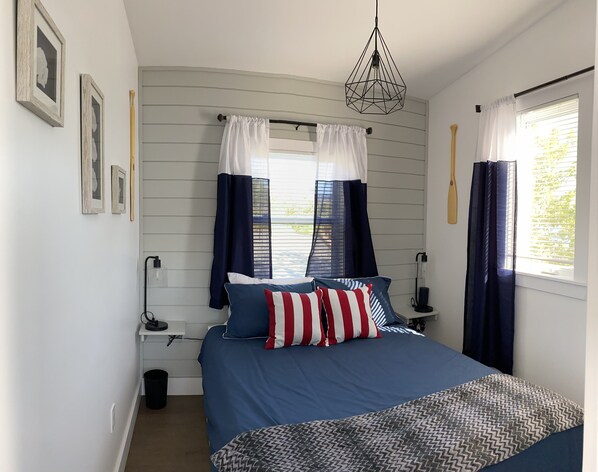 A bedroom with a bed, two bedside tables, a hanging light fixture, a window with curtains, and a wall-mounted shelf.
