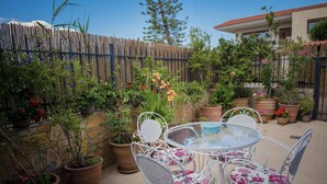 Un patio con un tavolo rotondo in vetro e sedie in metallo bianco con cuscini a motivi floreali, circondato da piante in vaso e un muro in pietra.