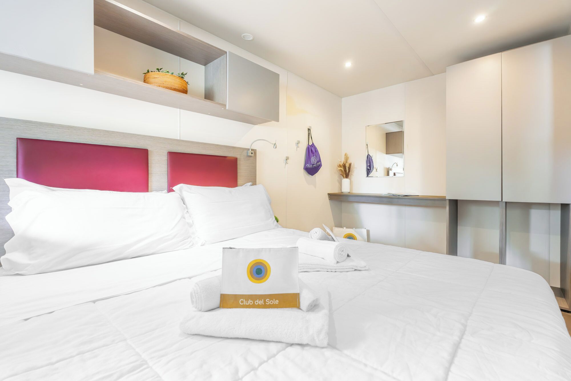 Habitación de hotel con cama, cabecero, estante con una cesta, espejo y toalla con un logotipo.