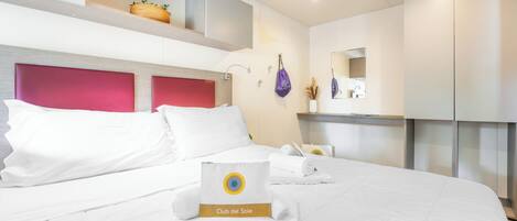 Habitación de hotel con cama, cabecero, estante con una cesta, espejo y toalla con un logotipo.