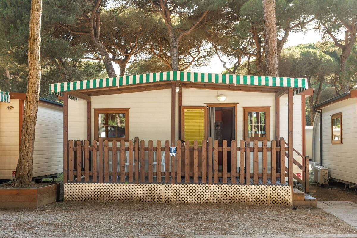 Eine kleine, eingeschossige Holzhütte mit Veranda, einem Holzzaun und einem Fenster mit gelbem Rollo.