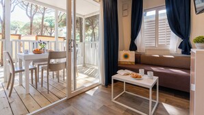 Una habitación acogedora con una puerta corrediza de vidrio que da a un espacio al aire libre, un sofá, una pequeña mesa con un libro y pasteles, y un televisor en la pared.