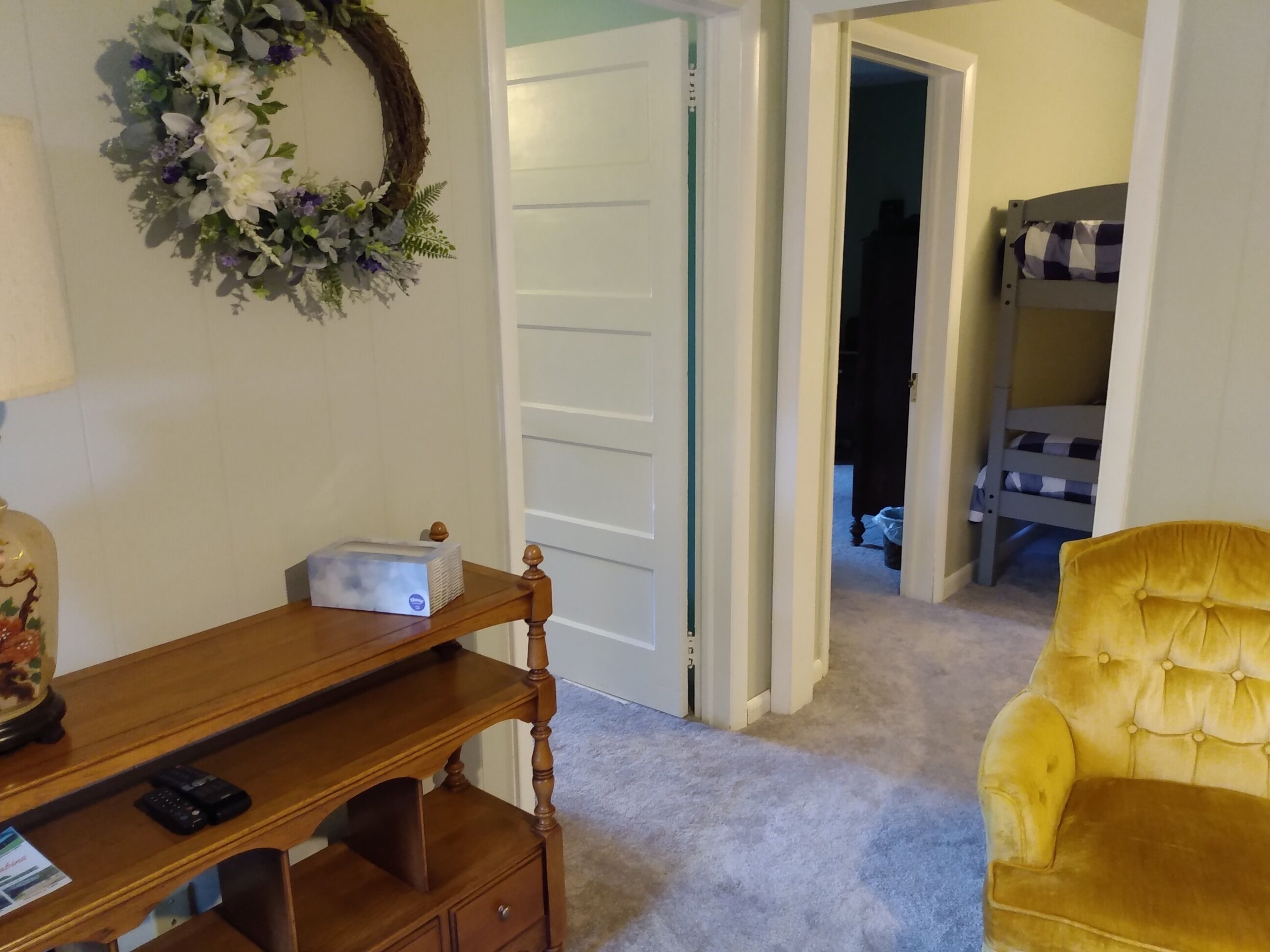 Ein Zimmer mit einem Holztisch, einem gelben Sessel, einer weißen Tür und einem Stockbett im Hintergrund.