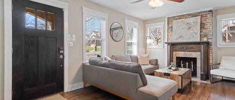 A living room with a dark wooden door, a grey sofa, a white armchair, a fireplace, and a ceiling fan.