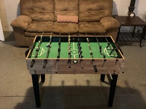 Une table de baby-foot dans une pièce où se trouvent un canapé marron et une petite table en arrière-plan.