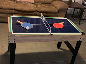 Mesa de ping pong con dos palas y una pelota en el centro, ubicada en una habitación con un sofá marrón y una mesita en el fondo.
