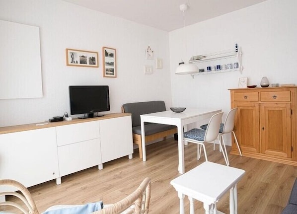 A modern, well-lit room with a white TV on a wooden cabinet, a grey sofa, a white dining table with chairs, and a wooden cabinet with shelves above. - Lüttjer Kiwitt - perfect location for short distances and relaxing days (Wangerooge)