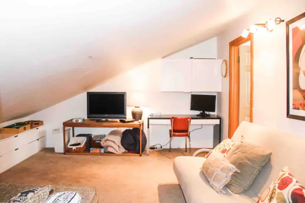 Quarto aconchegante e bem iluminado com televisão de tela plana, escrivaninha com computador, cadeira vermelha e sofá com almofadas estampadas.