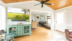 A spacious living room with a wooden ceiling, a ceiling fan, a large flat-screen TV mounted on the wall, a glass coffee table, a teal cabinet, and a brown leather sofa. - The Robin house - Close to Everything! (Mount Pleasant)