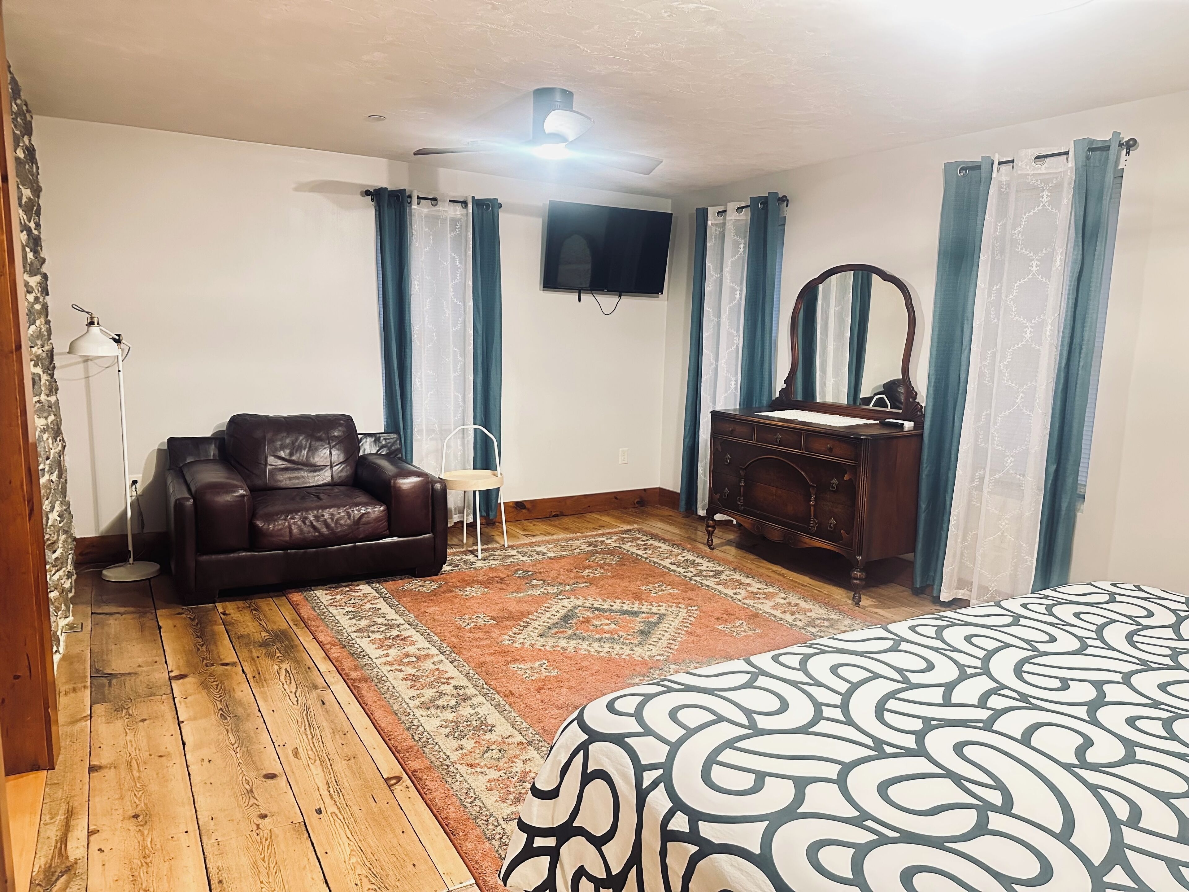 Una habitación con piso de madera, un sofá de cuero marrón, un aparador de madera, un espejo, un televisor en la pared y una colcha estampada.