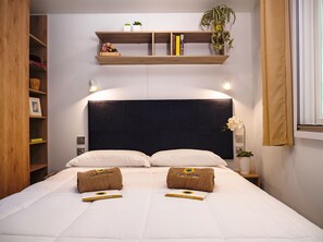 A hotel room with a bed, two pillows, a bedside table, a nightstand, a shelf with books and a plant, and a window with curtains. - Club Del Sole Marina Romea Easy Camping Village (Ravenna)