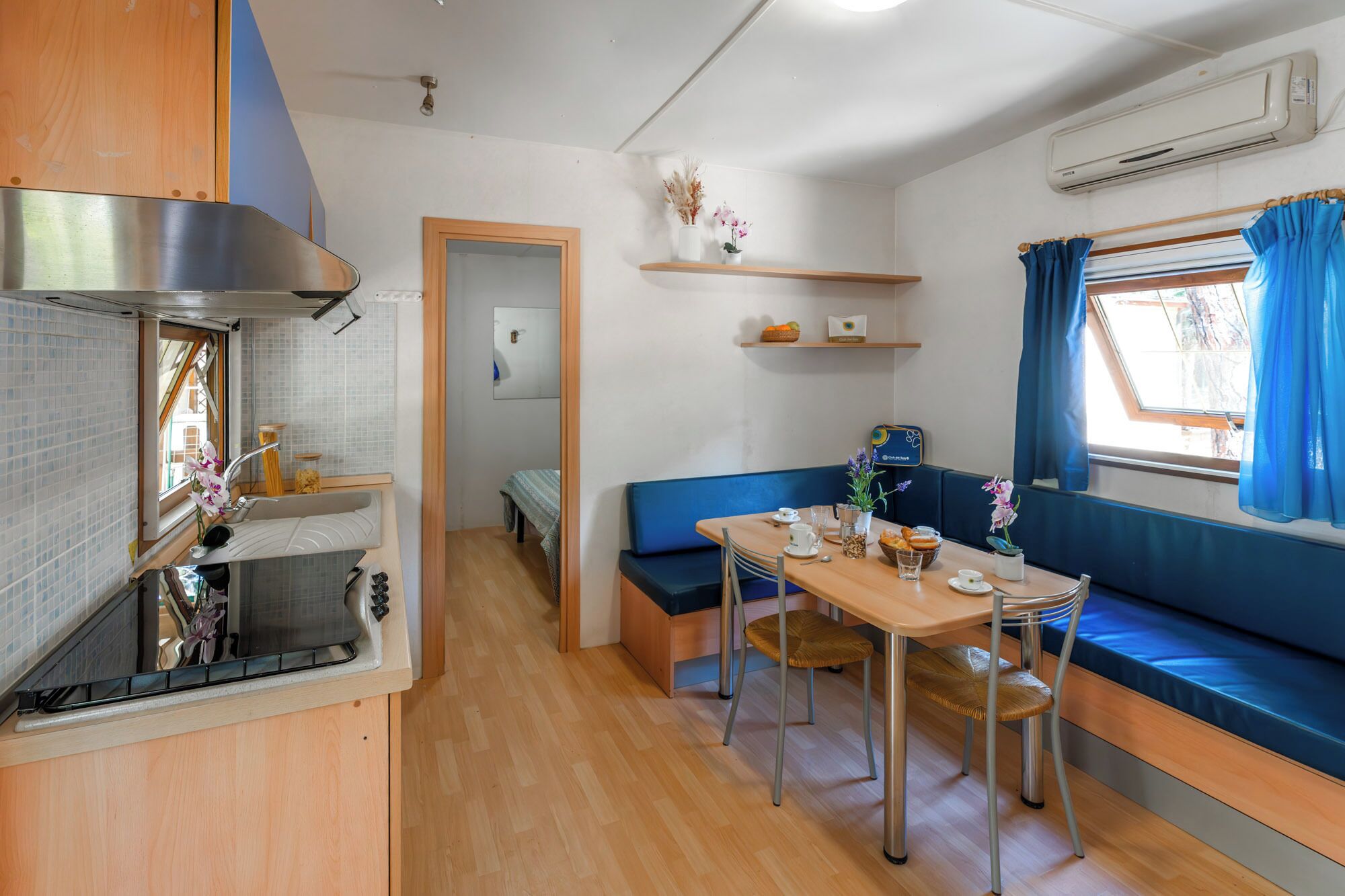 Une petite cuisine équipée avec une cuisinière, un évier et une petite fenêtre. Un coin repas, avec une table et des chaises, est situé à côté d’un banc bleu rembourré.