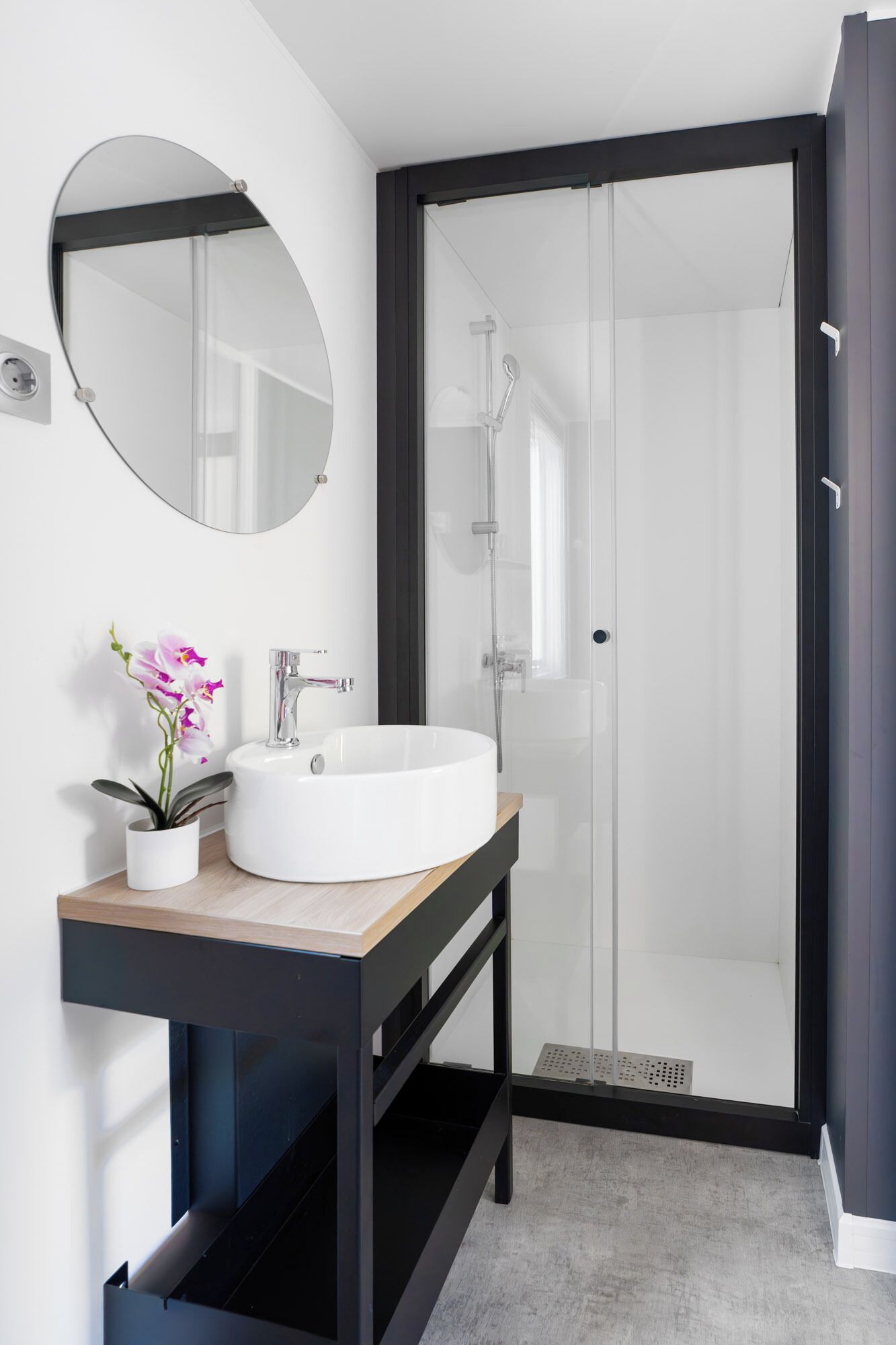 Une salle de bain moderne avec un miroir rond, un lavabo blanc et une cabine de douche en verre.