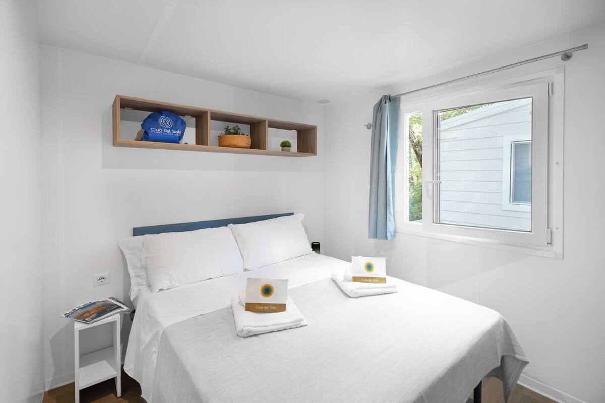 A bedroom with a bed, a nightstand, a shelf with items, and a window.