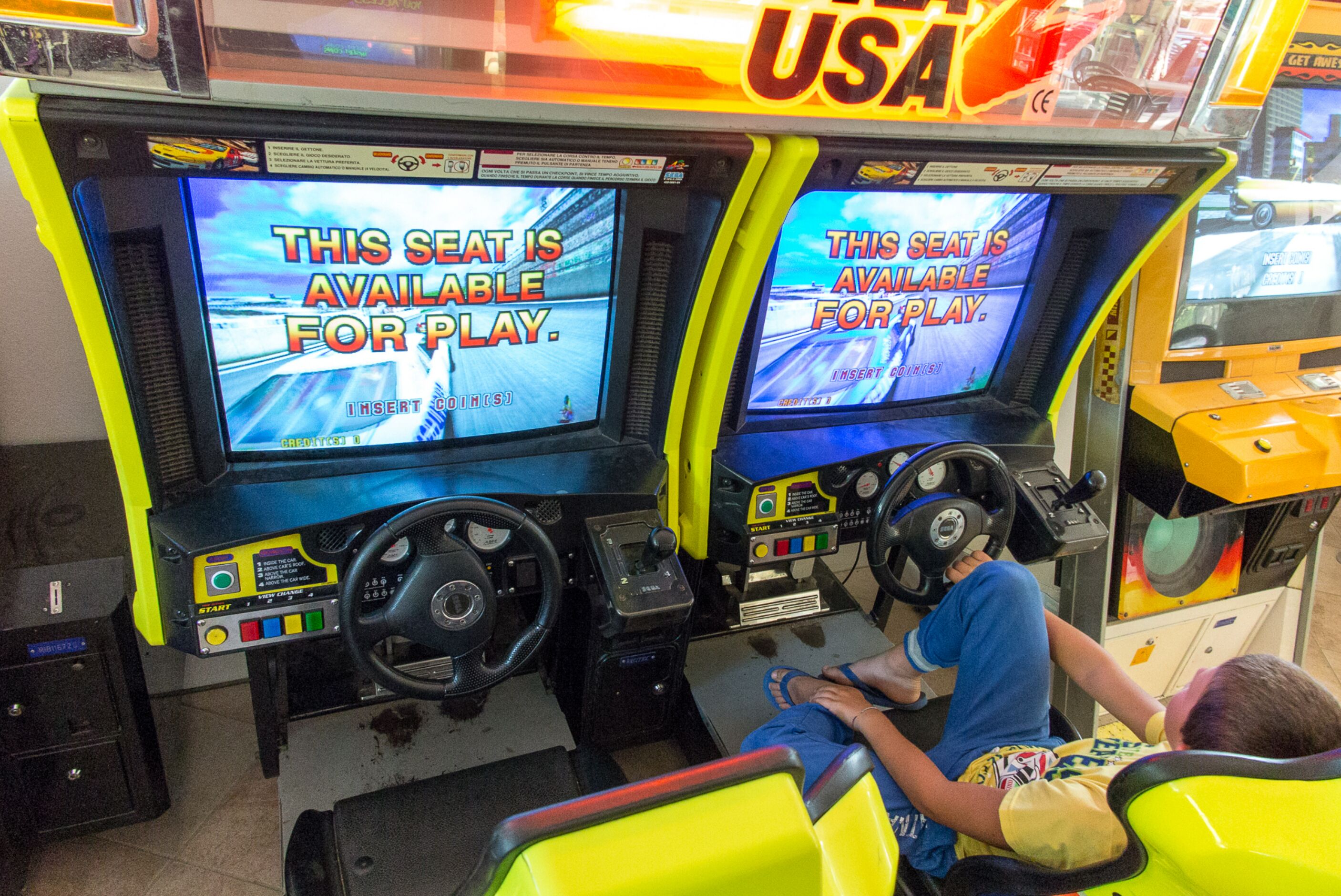 A video game arcade with two racing game machines, each with a steering wheel and a screen displaying a message about seat availability.