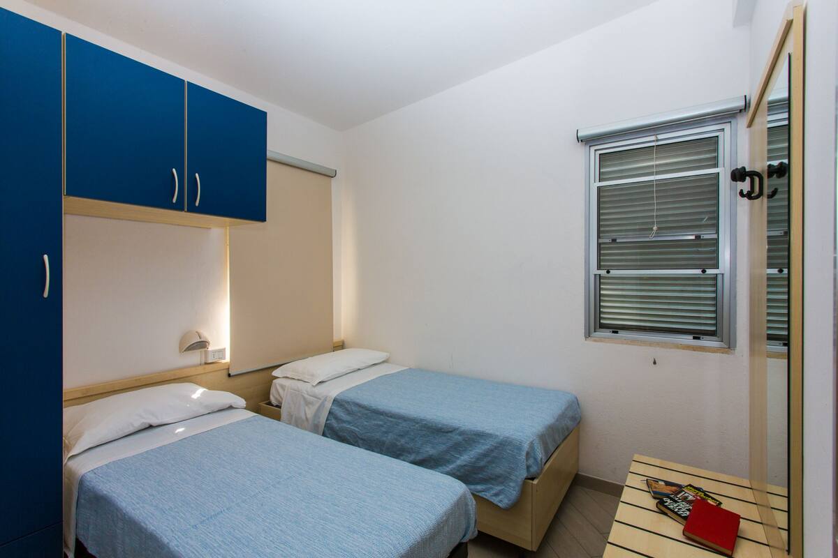 A small, tidy room with two single beds, a wooden table, a window with blinds, and a mirror.