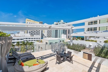A rooftop patio with lounge chairs, a seating area, a small outdoor kitchenette, and city views on a sunny day, surrounded by modern high-rise buildings.