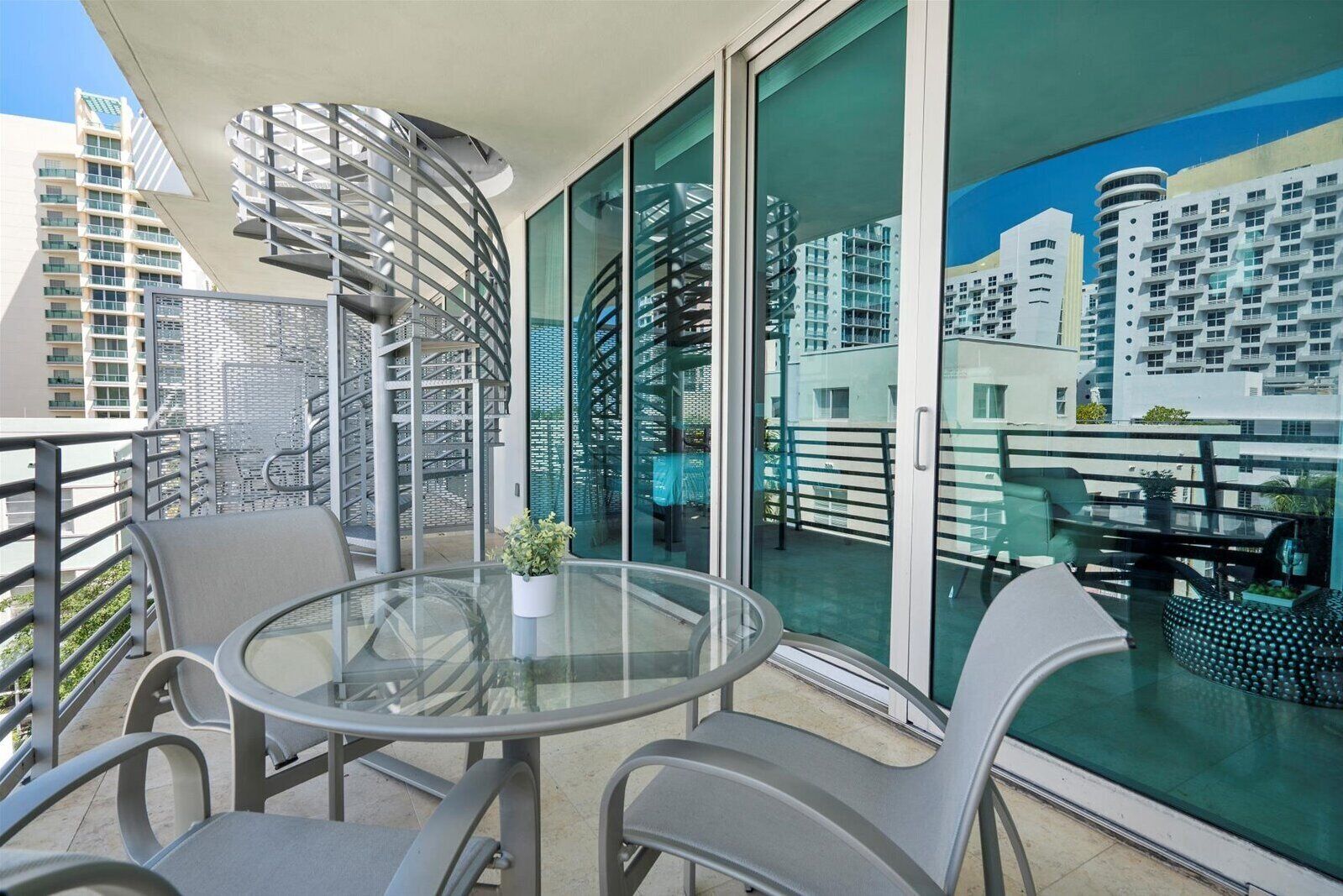 Un balcón con una mesa y sillas de vidrio, una planta en maceta y vistas a edificios modernos.