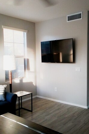 A living room with a flat-screen TV mounted on the wall, a window with blinds, a lamp, and a sofa.